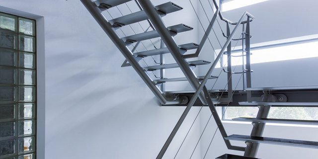 Eine moderne Treppe aus Metall mit schlichten Stufen, die in einen hellen Raum führt, beleuchtet von großen Fenstern.
