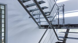 Eine moderne Treppe aus Metall mit schlichten Stufen, die in einen hellen Raum führt, beleuchtet von großen Fenstern.